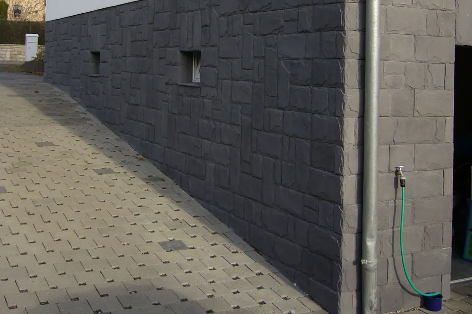Dark stone-pattern finish on the lower wall of a house beside paving stones